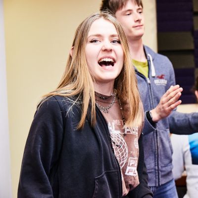 A girl wearing a brown t-shirt with shoulder length brown hair sings open mouthed. A boy plays the tambourine in the background.